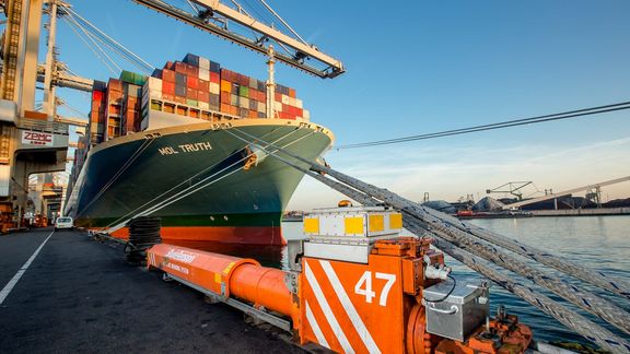 Grote containerboot met de naam "MOL TRUTH" ligt aangemeerd aan een kade in een haven. Het schip is beladen met kleurrijke containers en vastgemaakt met dikke trossen. Op de voorgrond staat een oranje en witte kadebescherming met het nummer 47. Op de achtergrond zijn havenkranen en industriële installaties zichtbaar onder een heldere blauwe lucht.erschip aangemeerd in een haven