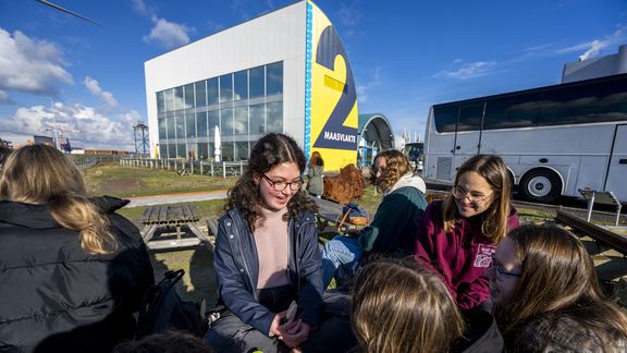 Een groep mensen zit buiten op houten picknickbanken bij een groot modern gebouw met een opvallend geel-blauw logo waarop “2 Maasvlakte” staat. Op de achtergrond staat een witte touringcar en is een open terrein zichtbaar onder een heldere blauwe lucht met enkele wolken.