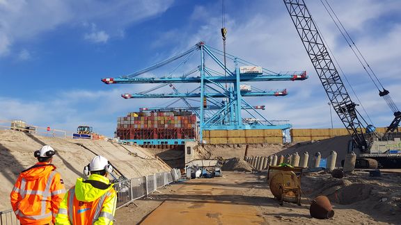 Een bouwplaats in een havengebied met grote blauwe containerkranen op de achtergrond en gestapelde containers. Op de voorgrond zijn twee personen in felgekleurde veiligheidsjassen en helmen te zien, die naar de werkzaamheden kijken. Er zijn zandheuvels, bouwmachines, een hijskraan en betonnen barrières zichtbaar, onder een heldere blauwe lucht met enkele wolken.