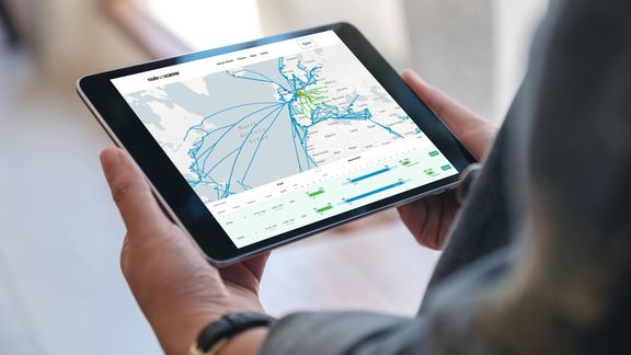 Een close-up van een tablet die een digitale kaart toont met scheepvaartroutes over Europa en de Atlantische Oceaan. Op het scherm zijn blauwe lijnen zichtbaar die de vaarroutes aangeven, groene markeringen voor havens en een gegevensoverzicht met grafieken en tabellen onder de kaart. De tablet wordt vastgehouden in een binnenomgeving met een neutrale achtergrond.