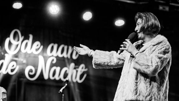 Zwart-witfoto van een persoon in een dikke bontjas die op een podium staat en spreekt in een microfoon, met een uitgestrekte arm richting het publiek. Op de achtergrond is een verlicht bord te zien met de tekst "Ode aan de Nacht" en enkele spotlights boven het podium
