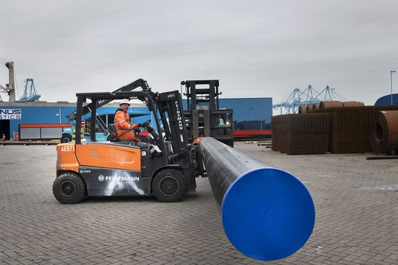 Een heftruck met oranje carrosserie en het logo van Peinemann op de zijkant verplaatst een grote metalen buis met een blauwe afgedichte uiteinde op een industrieterrein. Op de achtergrond zijn stapels metalen materialen en grote blauwe loodsen zichtbaar, evenals containerkranen in de verte. De ondergrond bestaat uit grijze bestrating en de lucht is bewolkt.
