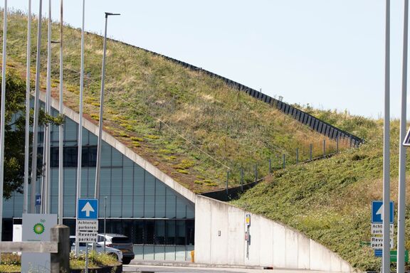 Een modern gebouw met een schuin dak dat volledig bedekt is met gras en vegetatie, zichtbaar vanaf de zijkant. Voor het gebouw bevindt zich een parkeerplaats met verkeersborden waarop “Achteruit parkeren / Reverse parking” staat. Links is een BP-logo zichtbaar op een bord. Op de achtergrond zijn enkele lantaarnpalen en een heldere blauwe lucht te zien. Het ontwerp van het gebouw benadrukt duurzaamheid en integratie met de natuurlijke omgeving.