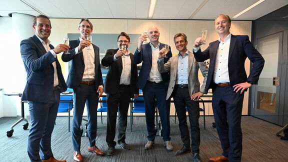 Zes personen in nette zakelijke kleding staan in een kantoorruimte en heffen glazen champagne voor een toast. Op de achtergrond zijn blauwe stoelen, een tafel en een wand met lichte panelen zichtbaar. De vloer is bedekt met donker tapijt en er is een glazen deur aan de rechterkant.