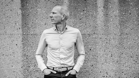 Eine Person steht vor einer groben Betonwand mit beiden Händen in den Hosentaschen. Die Person trägt ein helles, langärmeliges Hemd und eine dunkle Hose mit Gürtel. Die Aufnahme ist in Schwarz-Weiß gehalten, wodurch die Struktur der Wand und die Kleidung deutlich hervorgehoben werden.