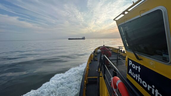 Een gele patrouilleboot van de havenautoriteit vaart over rustig water, waarbij een witte schuimstreep achter de boot zichtbaar is. Op de zijkant van de cabine staat de tekst “Port Authority”. In de verte is een groot vrachtschip te zien onder een licht bewolkte lucht met strepen van zonlicht, wat een serene maritieme sfeer creëert.