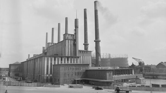 Zwart-witfoto van een groot industrieel fabriekspand met meerdere hoge schoorstenen die rook uitstoten. Het gebouw heeft een robuuste constructie met verticale lijnen en meerdere verdiepingen. Op de voorgrond is een open terrein zichtbaar met enkele voertuigen en een omheining. Op de achtergrond staan extra industriële structuren, waaronder een kraan en ronde opslagtanks. De lucht is deels bewolkt, wat de historische en industriële sfeer benadrukt.