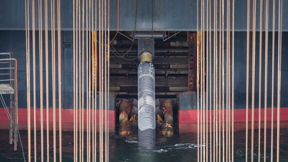 Close-up van een grote pijpleiding die door een opening in een offshore constructie wordt geleid. De pijp is zwart met markeringen en wordt ondersteund door zware mechanische klemmen. Verticale kabels of hijsdraden hangen aan weerszijden van de pijp. Het water is zichtbaar onderaan de constructie, en delen van het platform zijn rood en grijs gekleurd, wat wijst op een industriële maritieme omgeving.