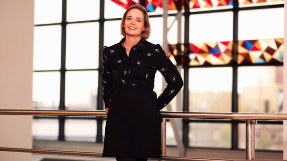 Een persoon staat in een moderne binnenruimte, leunend tegen een metalen railing. De persoon draagt een zwarte blouse met een subtiel patroon en een zwarte rok. Op de achtergrond zijn grote ramen zichtbaar met geometrische glas-in-loodpanelen in rood, geel en blauw tinten, waardoor een kleurrijk lichtspel ontstaat. Buiten is een stedelijk landschap vaag zichtbaar.