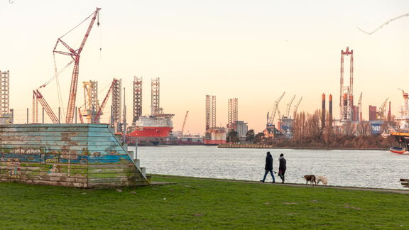 Mensen wandelen langs de waterkant met uitzicht op de Rotterdamse haven en industriele kranen.