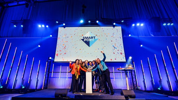 Een groep mensen staat op een podium tijdens een SmartPort-evenement. Op de achtergrond is een groot scherm zichtbaar met het SmartPort-logo en kleurrijke confetti. Het podium is verlicht met blauwe en paarse lampen, en er staan verticale lichtstaven langs de achterwand. Op een tafel rechts van het podium liggen enkele objecten, waaronder een laptop. De sfeer is feestelijk en professioneel, passend bij een officiële bijeenkomst of lancering.
