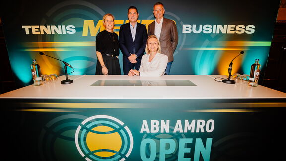 Vier personen staan achter een witte tafel met microfoons en waterflessen, in een ruimte met een groot achtergrondbord waarop de tekst “TENNIS MEETS BUSINESS” en “ABN AMRO OPEN” staat. Het decor bevat groene en gele accenten en een tennisbal-logo.