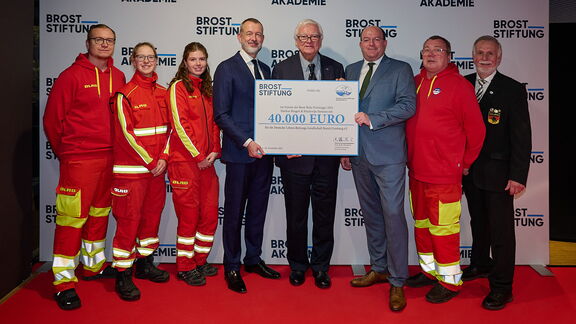 一群身穿正式西装和红黄色救援制服的人站在红地毯上，手持一张大型支票。支票上印有“BROST STIFTUNG”字样，金额为40.000欧元。背景是一块印有“BROST STIFTUNG”和“AKADEMIE”字样的宣传墙。