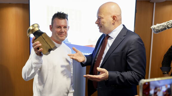 Twee personen staan in een ruimte met een houten wand en een scherm op de achtergrond. Eén persoon houdt een gouden beeldje vast, terwijl de andere persoon in een donker pak en stropdas een gesprek voert. Op de voorgrond is een microfoon en een camera zichtbaar, wat wijst op een formele prijsuitreiking of ceremonie