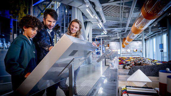 Drie personen bekijken een groot interactief scherm in een moderne tentoonstellingsruimte. De omgeving bevat glazen wanden, metalen constructies en een buisvormige installatie met oranje elementen aan het plafond. Rechts op de tafel liggen verschillende objecten en materialen, waaronder kleurrijke blokken en een piramidevormig model. De ruimte is helder verlicht en heeft een industriële uitstraling.