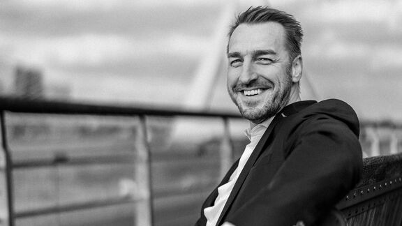 Een persoon in formele kleding zit op een houten bank naast een metalen railing, met een stedelijke achtergrond en een brug zichtbaar in de verte. De foto is in zwart-wit genomen.