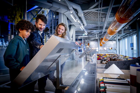 Drie personen bekijken een groot interactief scherm in een moderne tentoonstellingsruimte. De omgeving bevat glazen wanden, metalen constructies en een buisvormige installatie met oranje elementen aan het plafond. Rechts op de tafel liggen verschillende objecten en materialen, waaronder kleurrijke blokken en een piramidevormig model. De ruimte is helder verlicht en heeft een industriële uitstraling.