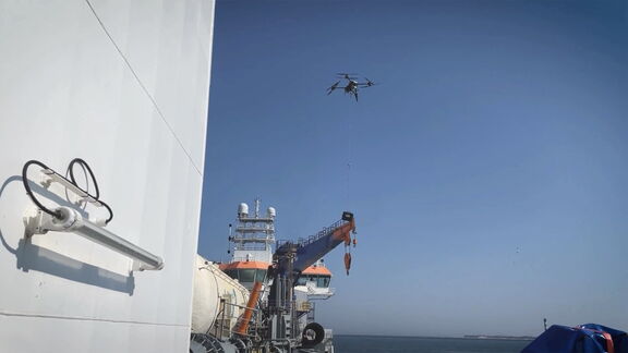 Een drone hangt boven een schip op open zee en laat een kabel zakken naar een kraan aan dek, met radarkoepels en antennes zichtbaar tegen een heldere blauwe lucht.