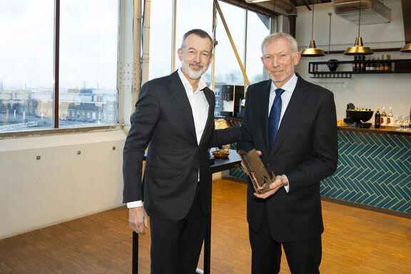 Twee personen in pakken staan binnen bij een bar, waarbij één persoon een award of trofee vasthoudt