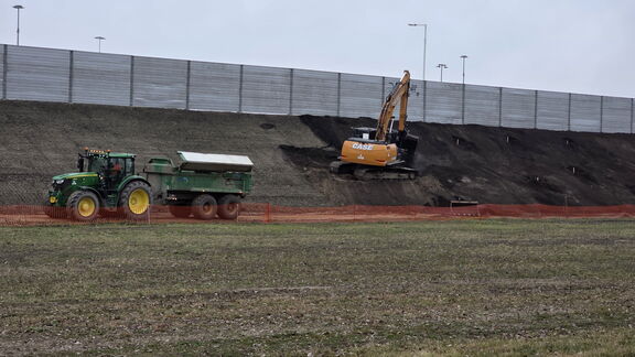 Graafmachine werkt op een verhoging naast een akker, met een tractor en aanhanger op de voorgrond
