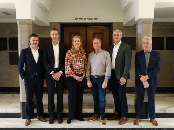 Zeven personen staan in een hal voor een deur met het bord ‘Commissariskamer