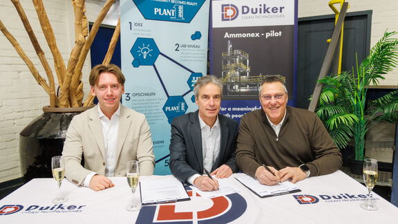 Drie personen zitten aan een tafel en ondertekenen documenten tijdens een officieel moment bij Duiker Clean Technologies, met banners en champagneglazen op de achtergrond.