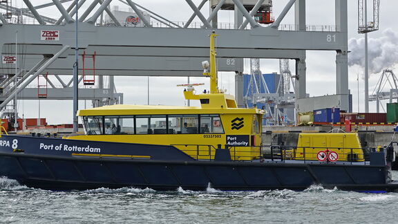 Geel-blauwe havenboot van Port of Rotterdam vaart langs containers.