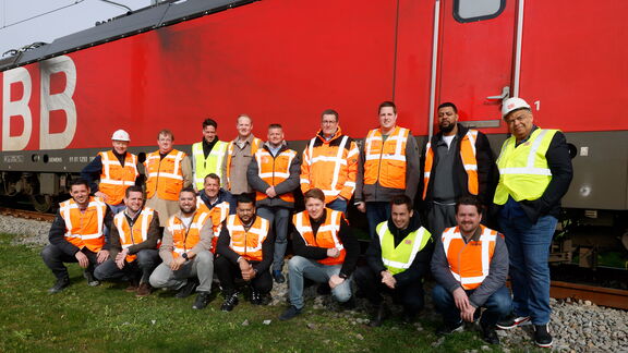 Group photo of about twenty people in orange and yellow safety vests posing in front of a red train wagon. They are standing on grass and gravel, some crouching and others standing, all looking at the camera