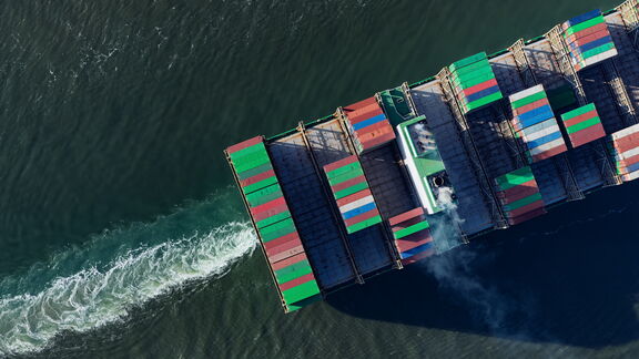 Luchtfoto van een groot vrachtschip dat over de oceaan vaart, beladen met kleurrijke containers in groen, rood en blauw, met een witte kielzog achter het schip en lichte rook uit de uitlaat.
