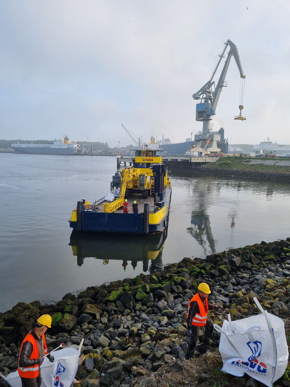 Werkboot en kraan in de haven terwijl medewerkers afval opruimen langs de oever.