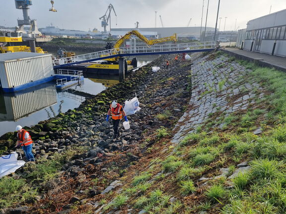 Werkboot in de haven terwijl medewerkers afval inzamelen langs de oever.