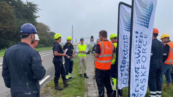 Groep wegwerkers met helmen en veiligheidsvesten staat langs de weg bij banners van Dutch Sweeper tijdens een briefing.