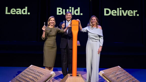 Drie personen houden symbolische sleutels omhoog naast een grote oranje sleutel, met de woorden “Lead. Build. Deliver.” op de achtergrond.