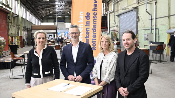 Vier personen staan bij een ondertekeningstafel in een industriële hal, met een oranje banner over werken in de Rotterdamse haven op de achtergron
