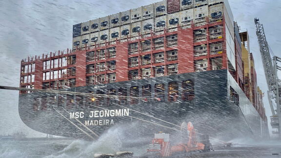 Containerschip MSC Jeongmin ligt aangemeerd aan de kade tijdens een zware storm, met opspattend water en oranje ShoreTension-units zichtbaar op de kade.
