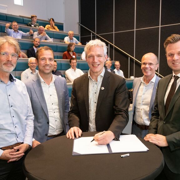 Een groep mensen staat rond een zwarte statafel in een vergaderruimte met tribune-opstelling. Op de tafel ligt een document dat wordt ondertekend. Op de achtergrond zitten meerdere personen op groene stoelen in oplopende rijen, alsof het een presentatie of officiële bijeenkomst betreft. De ruimte heeft witte muren en een donkere achterwand, met een trapleuning zichtbaar aan de rechterkant.