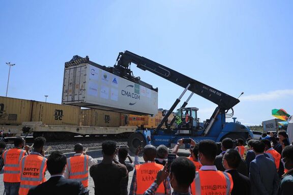Eine große blaue Reach-Stacker-Maschine hebt einen weißen Container mit der Aufschrift „CMA CGM“ auf einen Güterwaggon. Im Hintergrund sind mehrere gelbe MSC-Container auf Eisenbahnwaggons sichtbar. Im Vordergrund stehen zahlreiche Personen in orangefarbenen Sicherheitswesten mit Aufschriften wie „Safety Officer“ und „Ethiopian“, die den Vorgang beobachten. Der Himmel ist klar und blau, und die Szene zeigt eine typische Umschlagsituation im Güterverkehr.