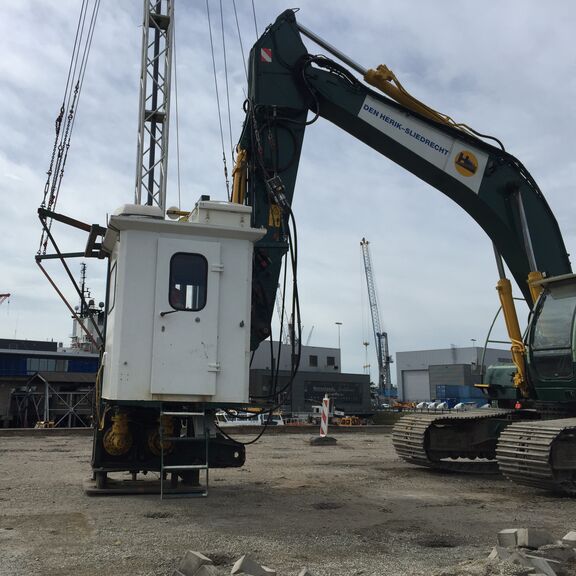 Een grote graafmachine met rupsbanden en een hijsinstallatie staat op een bouwterrein. Aan de arm van de machine hangt een witte cabine met ramen, bevestigd aan een metalen frame. Op de arm van de machine staat de tekst “DEN HERIK-SLIEDRECHT”. Op de achtergrond zijn industriële gebouwen, kranen en andere constructies zichtbaar, evenals een bewolkte lucht. De ondergrond bestaat uit zand en grind, typerend voor een industriële werkomgeving.