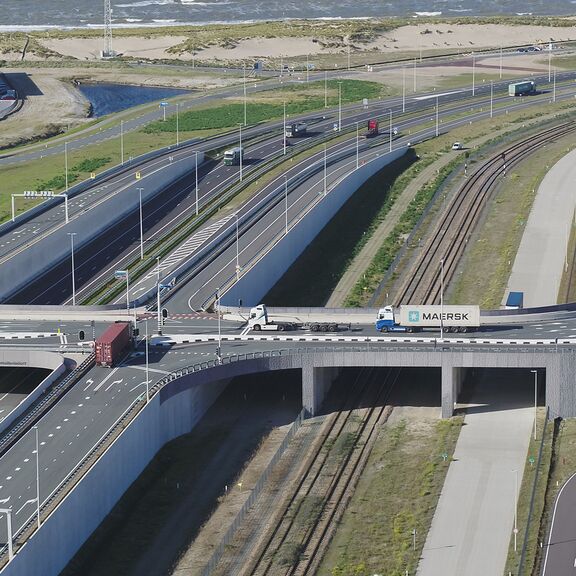 Luftaufnahme eines komplexen Straßennetzes mit mehreren Fahrspuren, Brücken und Kurven, kombiniert mit parallel verlaufenden Eisenbahngleisen. Auf den Straßen fahren Lkw, darunter ein Maersk-Container-Lkw, sowie andere Fahrzeuge. Im Hintergrund sind ein großes Industriegebäude, Container und Grünflächen sichtbar.