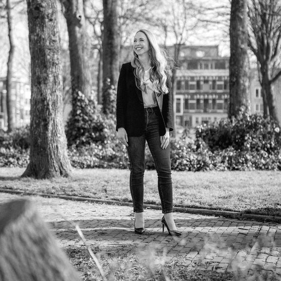 Black-and-white photo of an individual standing among tall trees in an outdoor environment. The person is wearing a dark blazer over a light top and fitted jeans, with long wavy hair visible. In the background, there are shrubs and multi-story buildings with large windows, suggesting an urban park or garden area.