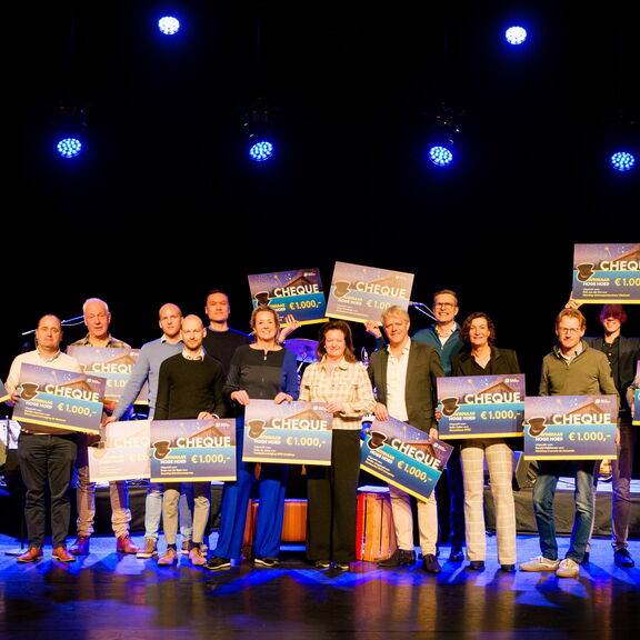Groep mensen op een podium tijdens een prijsuitreiking, elk houdt een grote cheque van €1.000 vast. Het podium is verlicht met blauwe spotlights en de achtergrond is donker, waardoor de cheques duidelijk zichtbaar zijn.