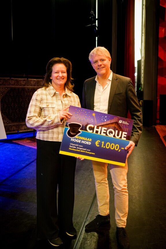 Twee personen staan op een podium en houden samen een grote cheque vast met een waarde van €1.000. Op de cheque staat de tekst “Winnaar Hoge Hoed” en er zijn feestelijke elementen zoals sterren en vuurwerk afgebeeld. De achtergrond toont een theateromgeving met rode gordijnen en donkere vloer.