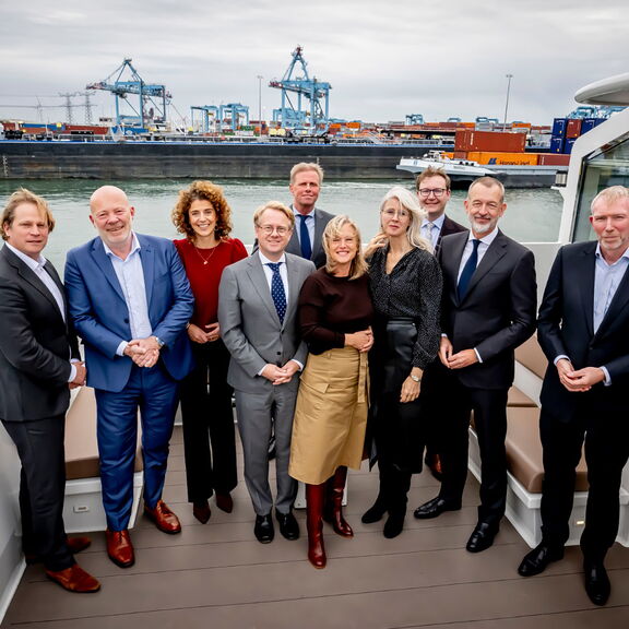 Een groep mensen staat samen op het dek van een boot in een havenomgeving. Op de achtergrond zijn containers, kranen en schepen zichtbaar, wat wijst op een industriële havenlocatie. De personen zijn netjes gekleed in zakelijke kleding, waaronder pakken en