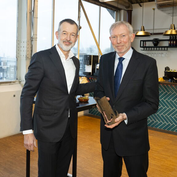 Twee personen in pakken staan binnen bij een bar, waarbij één persoon een award of trofee vasthoudt