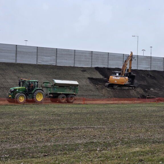 Graafmachine werkt aan een talud, terwijl een tractor met aanhanger over het terrein rijdt.