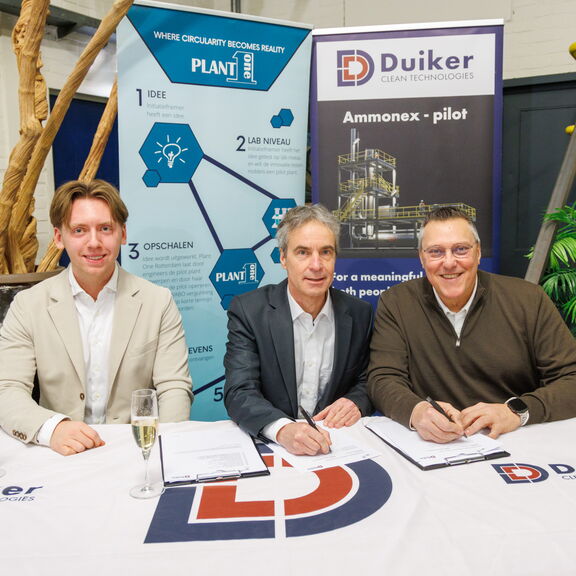 Drie personen zitten aan een tafel en ondertekenen documenten tijdens een officieel moment bij Duiker Clean Technologies, met banners en champagneglazen op de achtergrond.