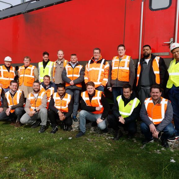 Groepsfoto van ongeveer twintig personen in oranje en gele veiligheidshesjes die poseren voor een rood treinwagon. Ze staan op gras en grind, sommige hurkend en anderen staand, allemaal kijkend naar de camera.