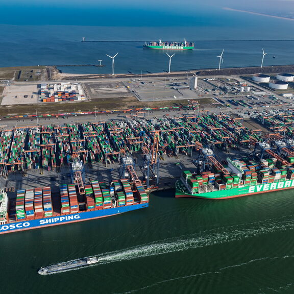 Luchtfoto van de RWG‑terminal met meerdere grote containerschepen, waaronder COSCO SHIPPING en EVERGREEN, aangemeerd aan de kade. Talloze kranen laden en lossen containers, terwijl op de achtergrond een uitgestrekt terrein met gestapelde containers en win