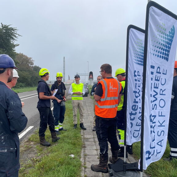 Groep wegwerkers met helmen en veiligheidsvesten staat langs de weg bij banners van Dutch Sweeper tijdens een briefing.