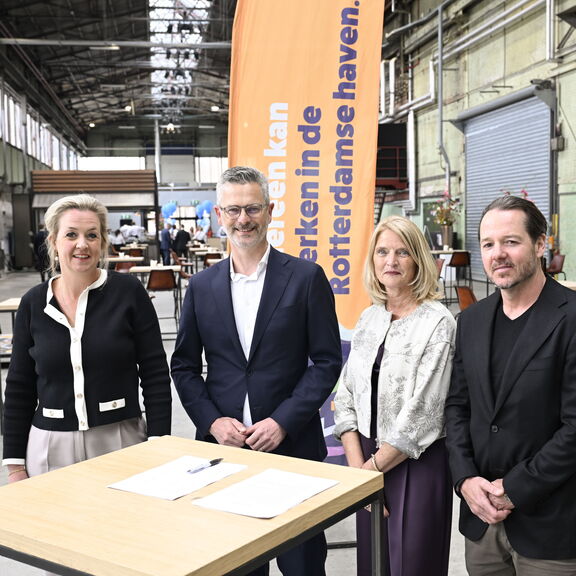 Vier personen staan bij een ondertekeningstafel in een industriële hal, met een oranje banner over werken in de Rotterdamse haven op de achtergrond.